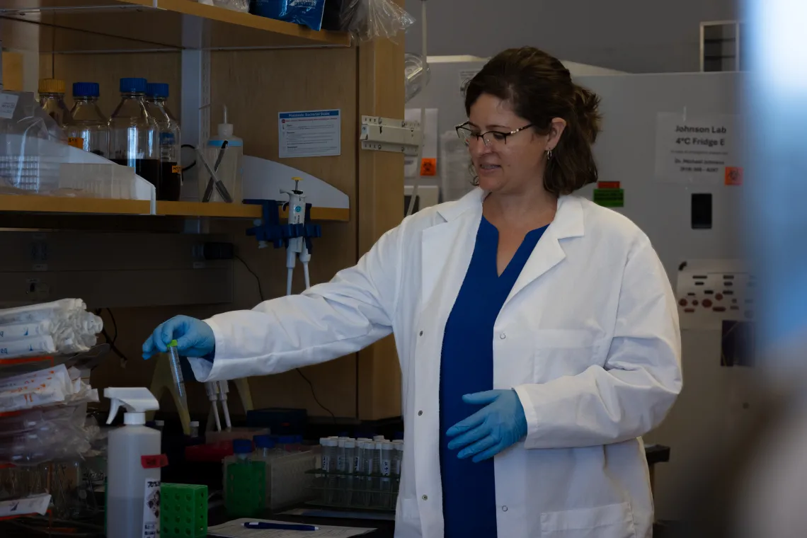 Person in a white coat and blue gloves holds a plastic vial in a laboratory