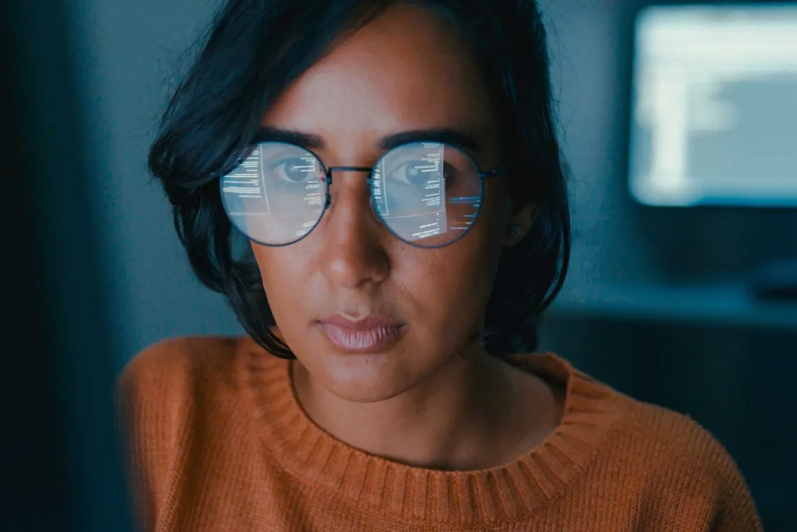 Person wearing glasses with computer screen reflections visible on the lenses, looking intently forward in a dimly-lit room.