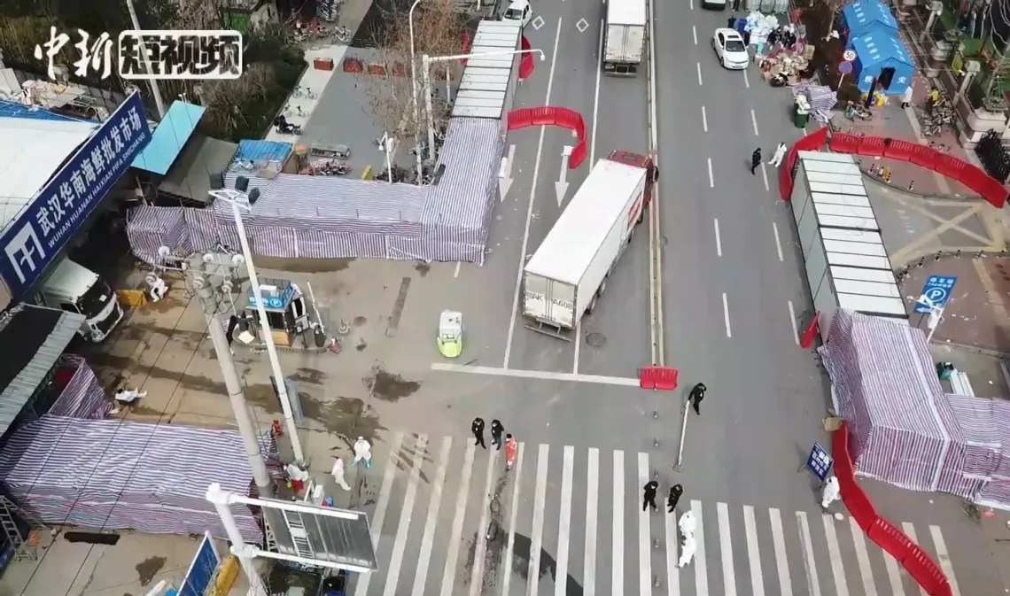 Aerial view of a busy street intersection with pedestrians crossing, surrounded by blue-roofed market stalls. Signage in Chinese characters is visible on nearby buildings.