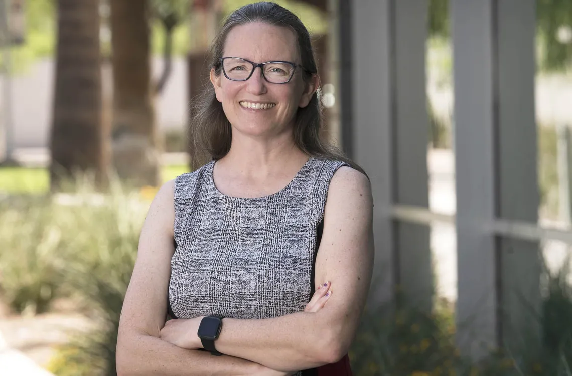 Person standing outdoors, smiling at the camera with arms crossed, wearing glasses, a gray sleeveless top, and a smartwatch.