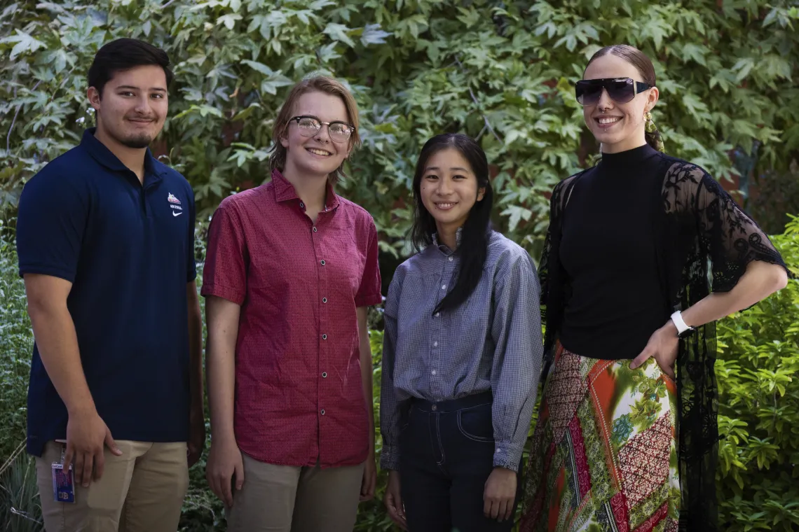 Four people smiling and standing together outdoors, with greenery in the background. From left to right, one person is wearing a blue polo shirt, the next is in a red button-down, followed by a person in a light blue shirt, and another in a black top with lace sleeves.