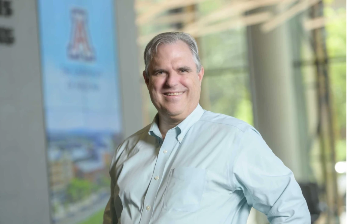 Individual smiling in front of a banner displaying 'The University of Texas at Arlington,' set against a light-filled, blurred background of a building interior.