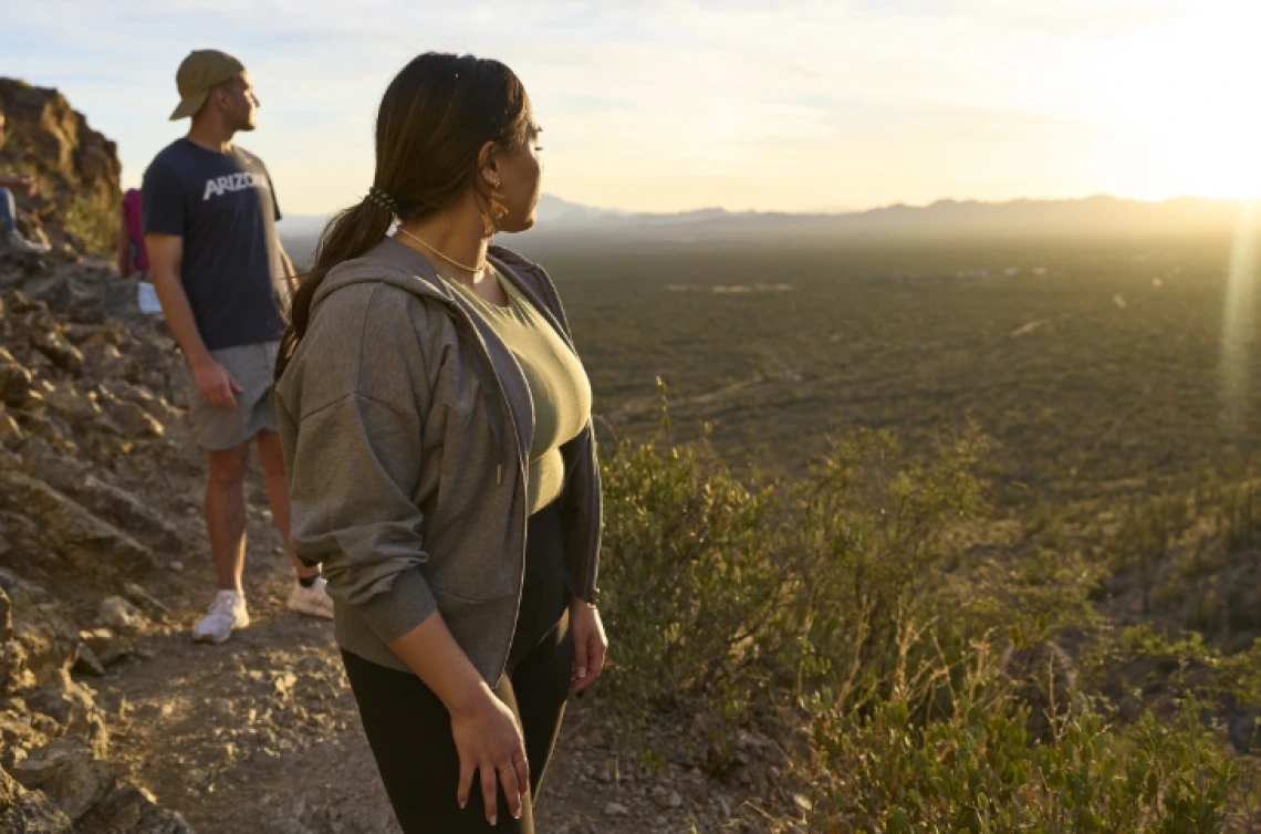 Two people standing on a rocky trail, overlooking a vast desert landscape at sunset. One individual is wearing a hat