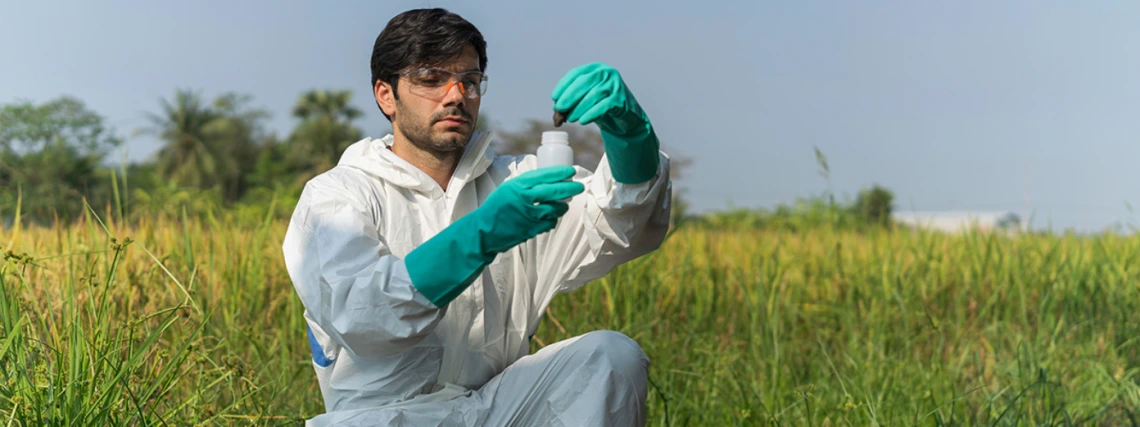 A scientist in a white lab coat and gloves examining a sample in a flask outdoors in a grassy field.