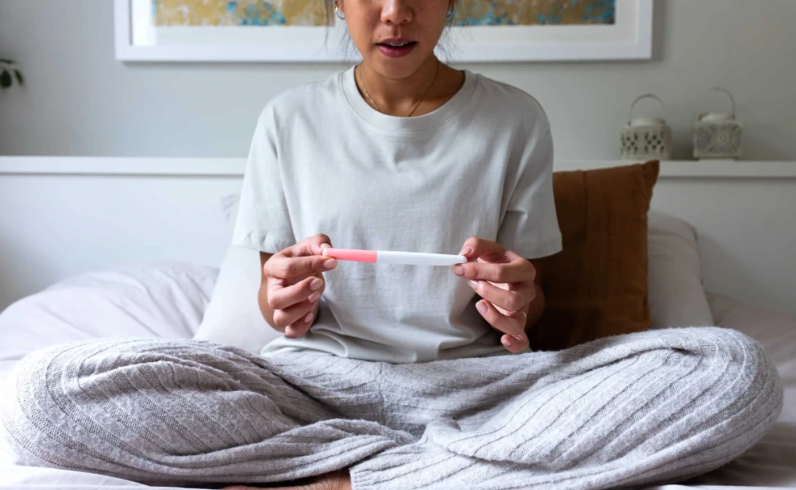 Person sitting on a bed, holding a pregnancy test, with a focused expression.