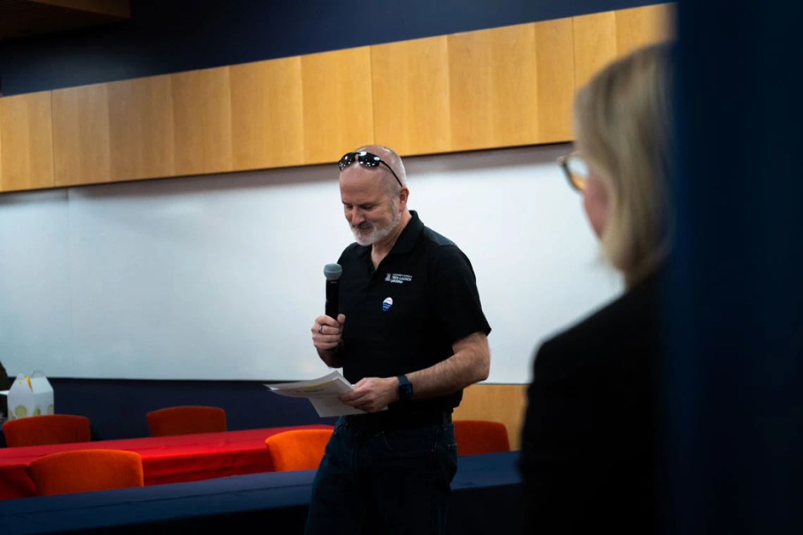 A person speaking into a microphone while holding papers, standing in front of an audience in a room with blue and orange chairs. Another person is visible in the foreground on the right side.