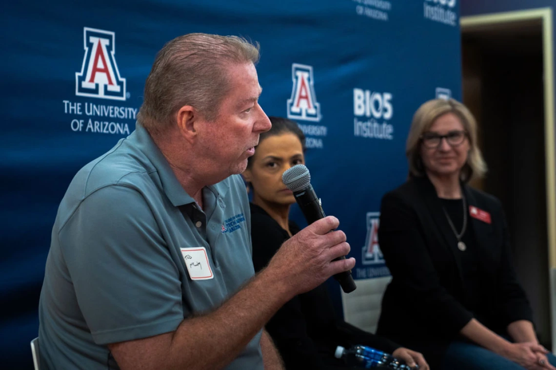 Three panelists at a University of Arizona event, one speaking into a microphone. The backdrop features the university and BIOS Institute logos.