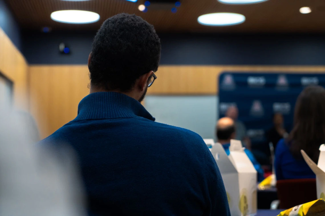 Person with curly hair sitting in a conference room, viewed from behind, focusing on a presentation at the front.