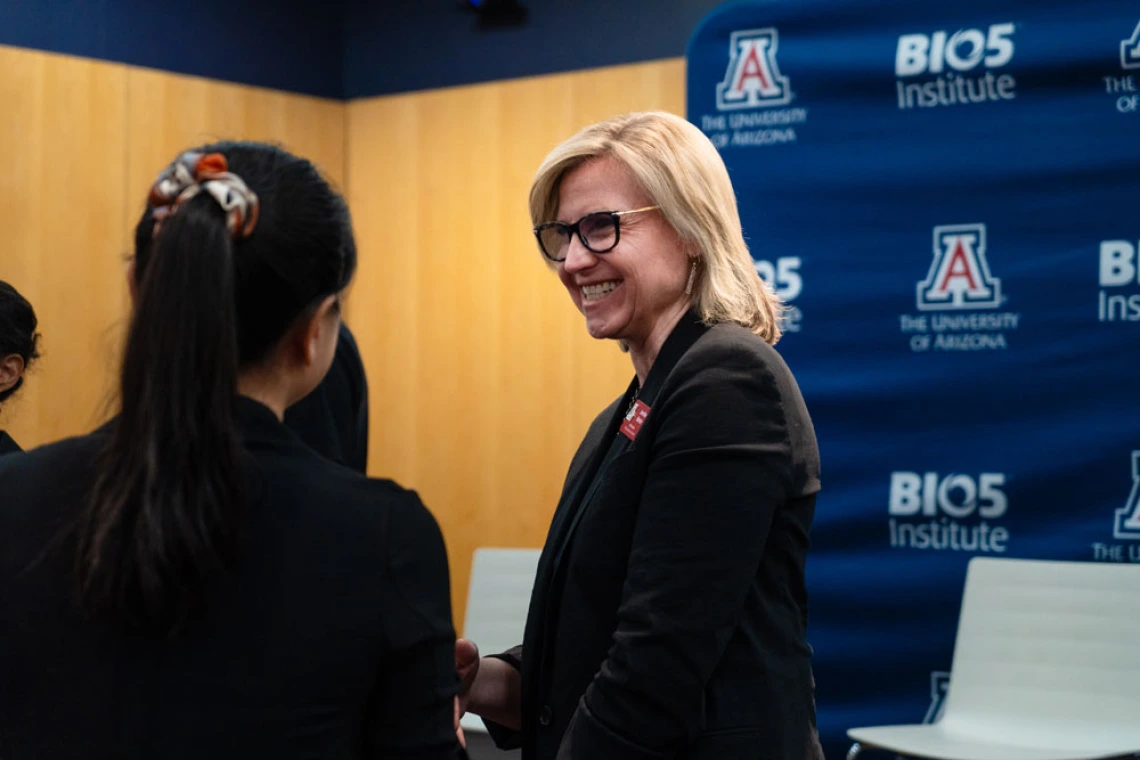 Two individuals engaging in conversation at a University of Arizona BIO5 Institute event. One person, seen from the back, has shoulder-length hair tied with a patterned scarf, while the other, facing forward, is smiling and has short blonde hair, wearing glasses and a name tag. The background features banners with the University of Arizona and BIO5 Institute logos.