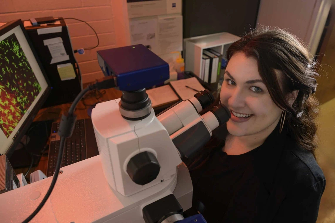 Person smiling at camera while working with a microscope in a laboratory.