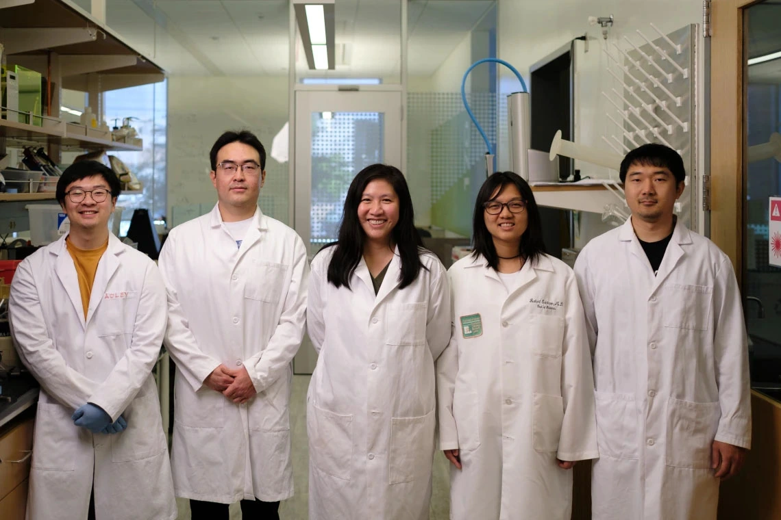 Five laboratory technicians wearing white lab coats are standing in a research lab. Each individual is smiling and facing the camera.