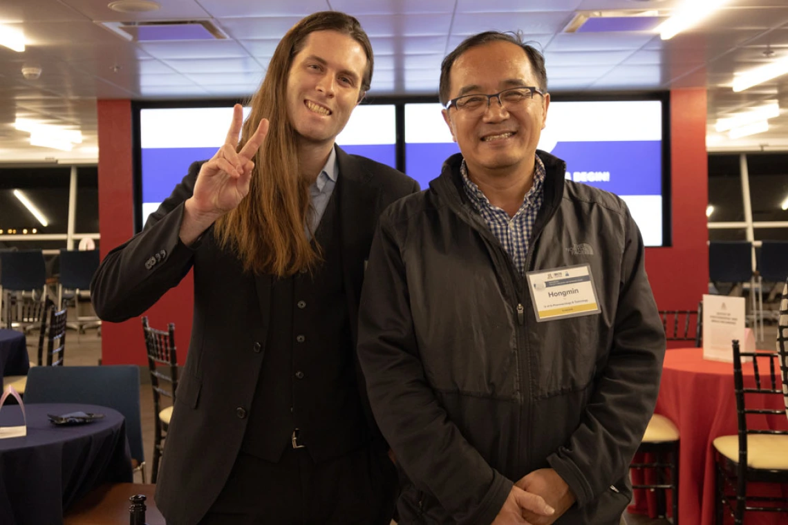 Two individuals at a professional event, one making a peace sign, with name tags visible and a conference background.