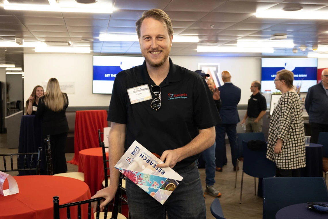 Person smiling at the camera, holding a brochure and wearing a badge, standing in a busy event space with people in the background.