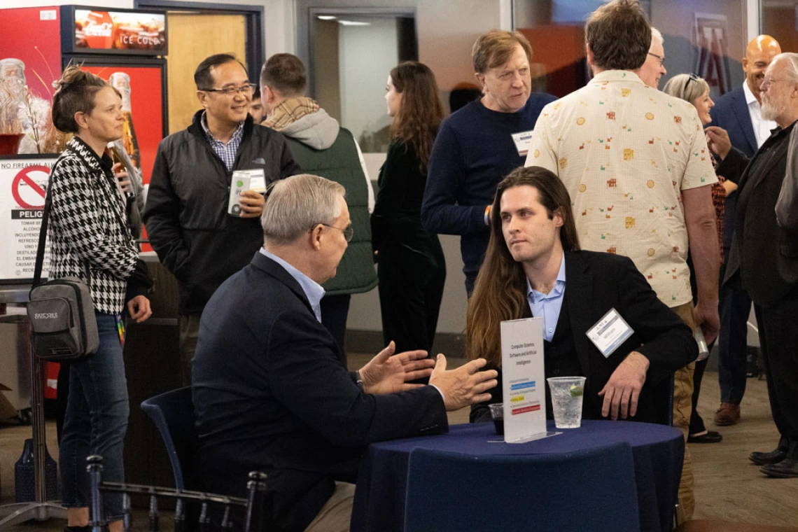 A networking event at a conference with attendees engaging in discussions. Two sit at a table and other individuals converse in the background near refreshment stands.