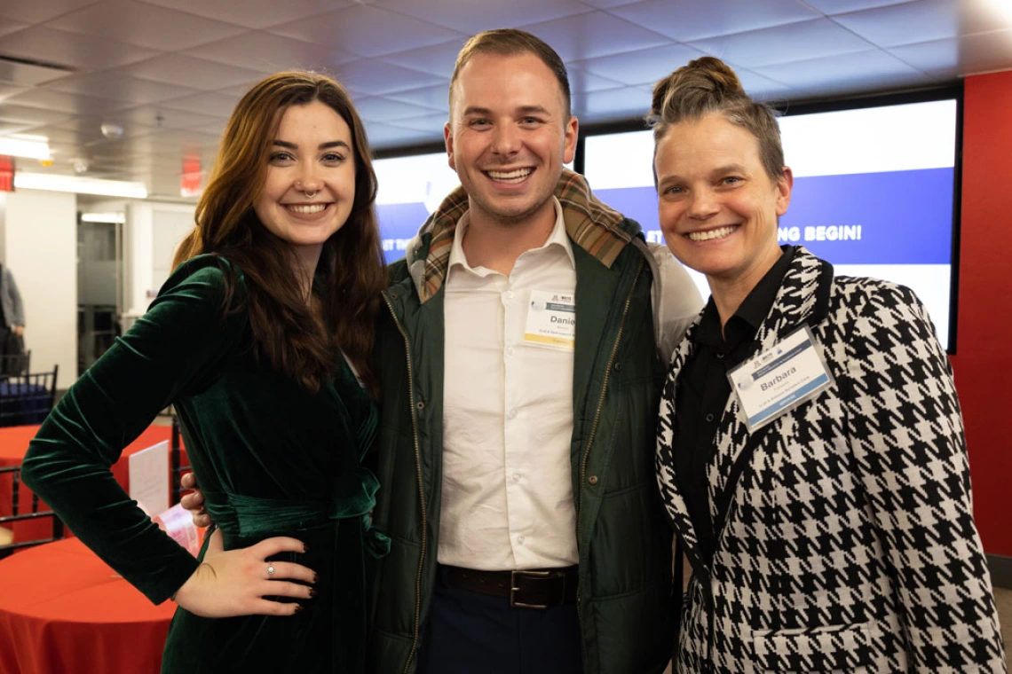 Three individuals smiling at a networking event, standing in front of a presentation screen that displays text.