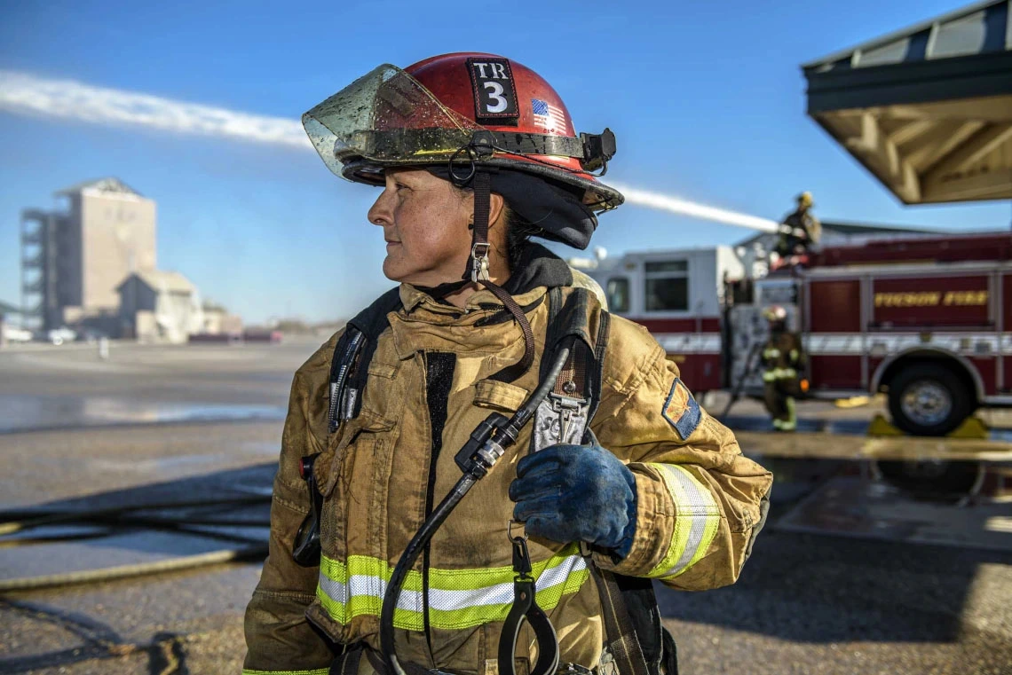 A firefighter in full gear stands in front of a fire truck labeled "Fresno Fire," with industrial buildings in the background. The firefighter is holding a helmet under the left arm and a radio in the gloved right hand.