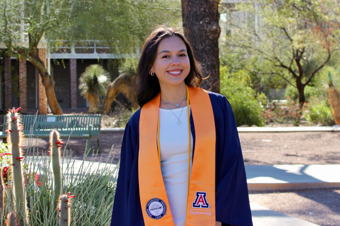 A person wearing a University of Arizona engineering graduation stole stands smiling outdoors in a sunny campus setting.