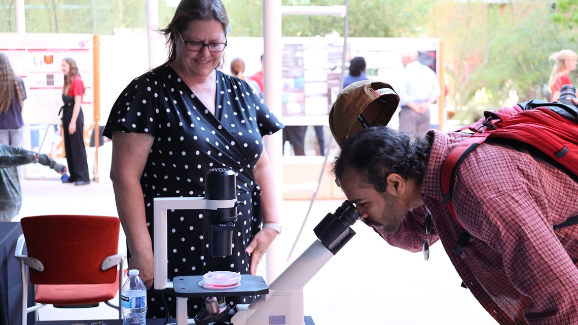 Person looks into a microscope while a researcher smiles and looks on