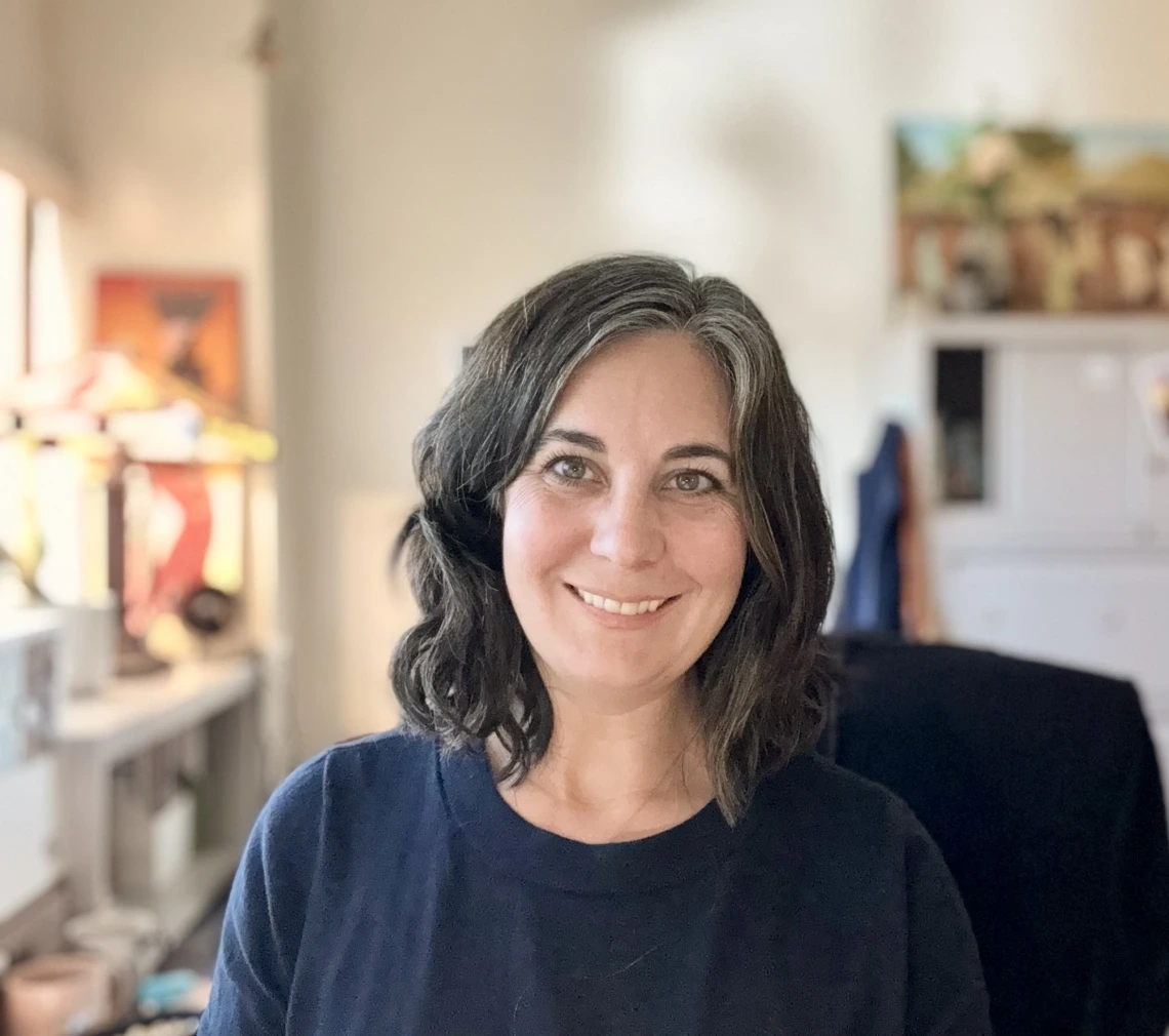 Portrait of a person smiling, with shoulder-length curly hair, wearing a blue top, in a well-lit room featuring art supplies and paintings in the background.