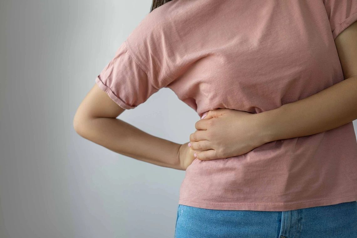 Person in a pink shirt holding their lower back, possibly indicating back pain, against a plain grey background.