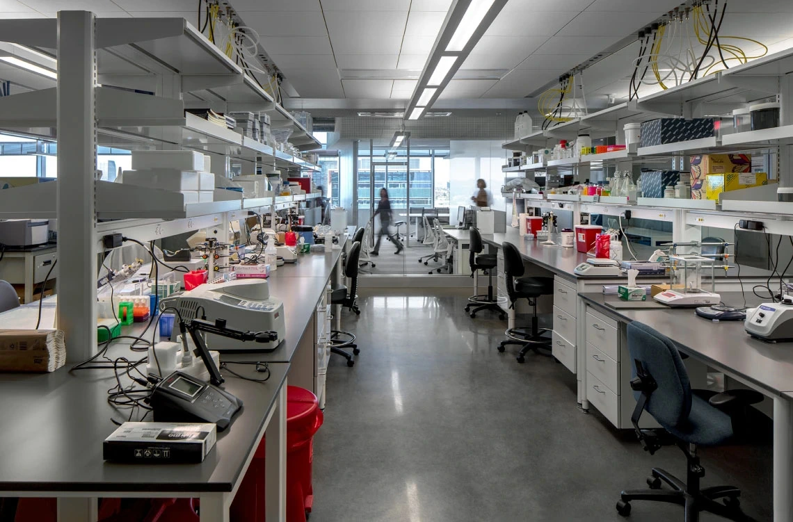 Interior of a modern laboratory with multiple workstations equipped with scientific instruments, computers, and laboratory supplies. Several chairs are positioned around the benches. Bright lighting and organized shelves are visible.