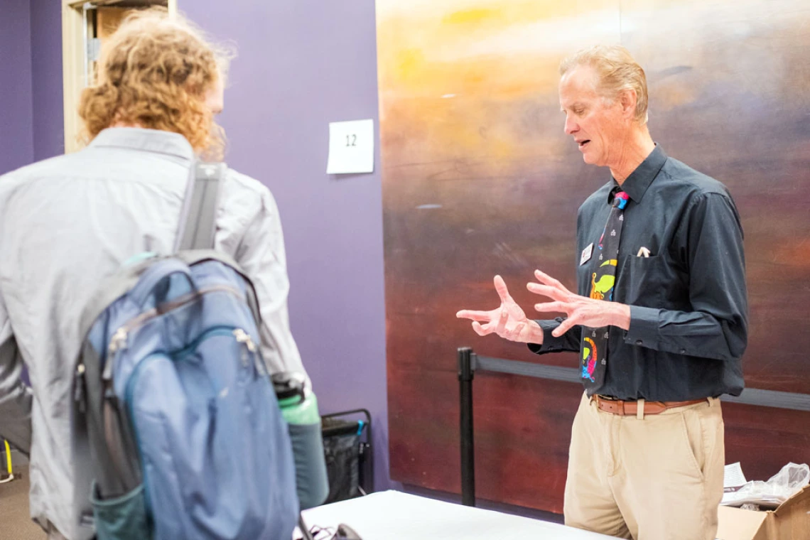 Two people converse, with one person gesturing expressively while the other listens with a backpack on. A large abstract painting hangs in the background.
