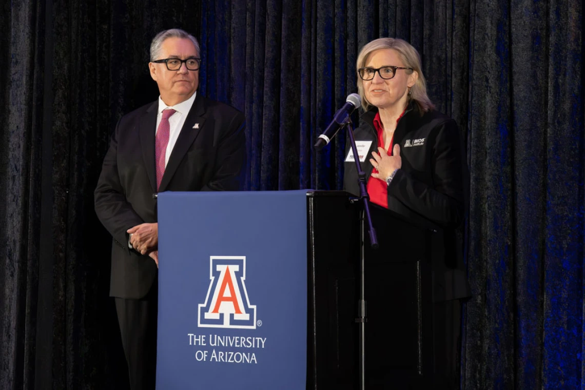 Two individuals stand at a podium with the University of Arizona logo, one speaking and the other listening attentively.