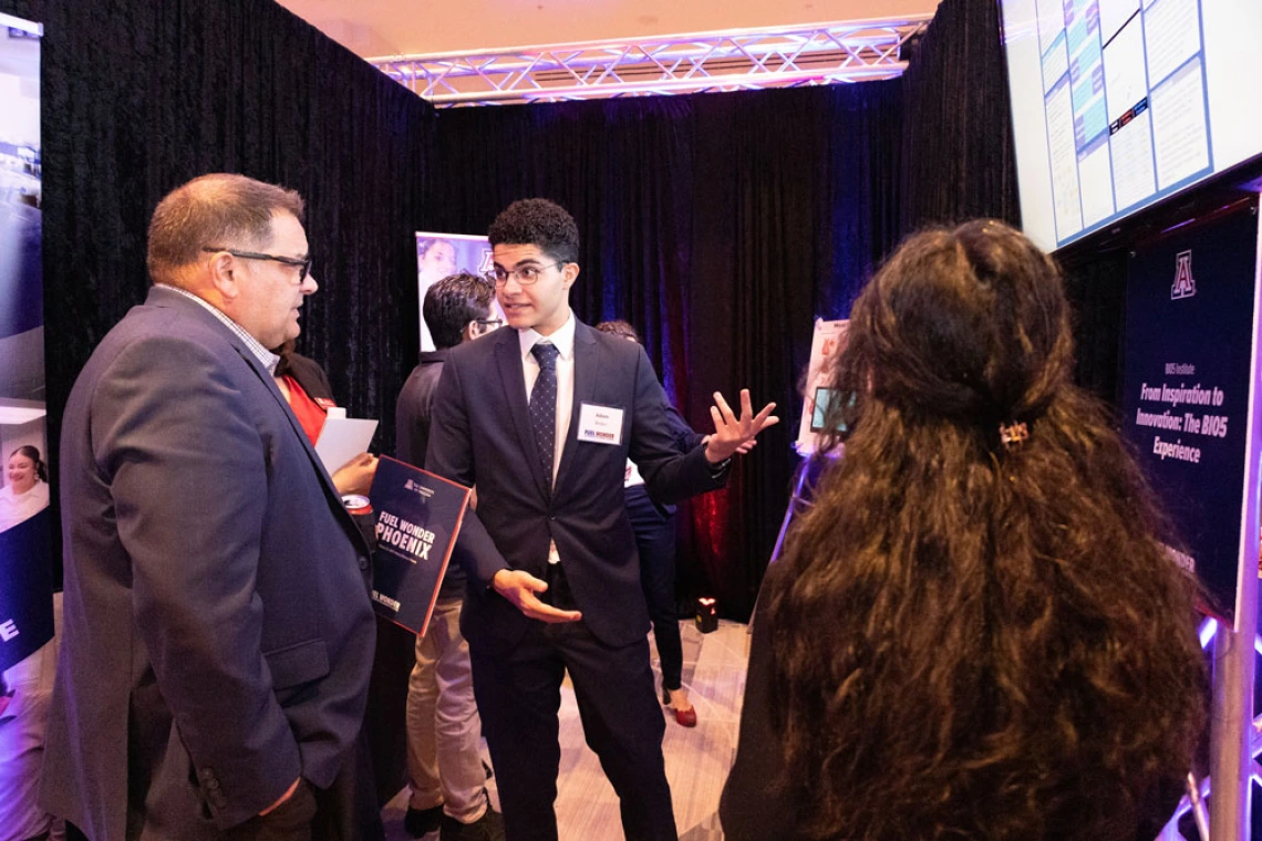 A person engaging in a discussion with others at a professional event hosted by the University of Arizona BIO5 Institute, standing beside a display with information about innovation and technology.