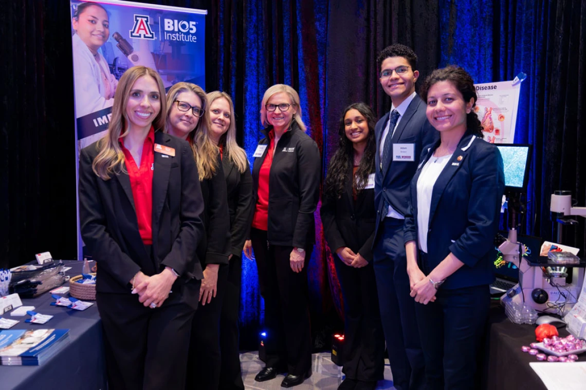 Group of seven individuals standing with confident smiles at a University of Arizona BIOS Institute recruitment booth, where banners and promotional materials are visible in the background.