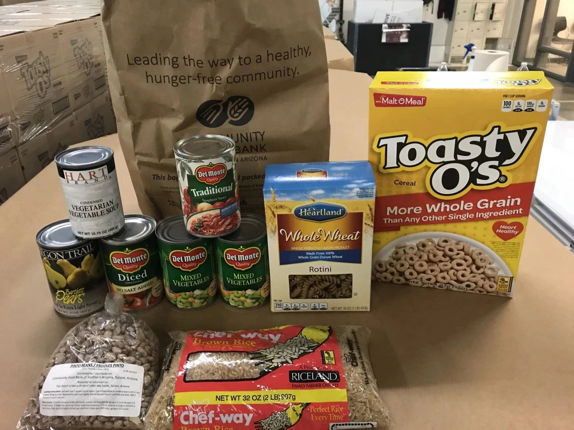 Assortment of food items for donation, including cans of vegetables, whole wheat pasta, a box of Toasty O's cereal, and a bag of rice, displayed on a table with a paper bag labeled "Leading the way to a healthy, hunger-free community.