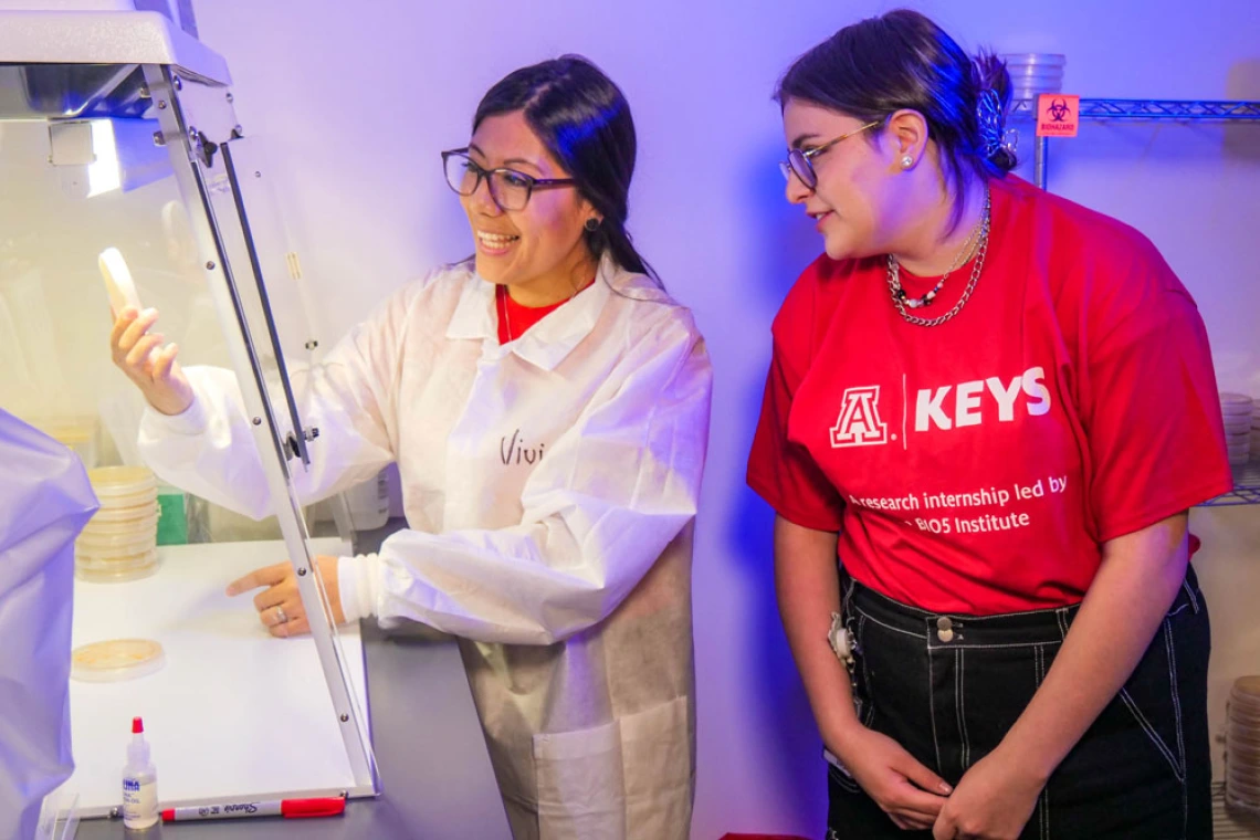 Person in white lab coat holds up petri dish underneath a hood while a person in a red KEYS t-shirt looks on