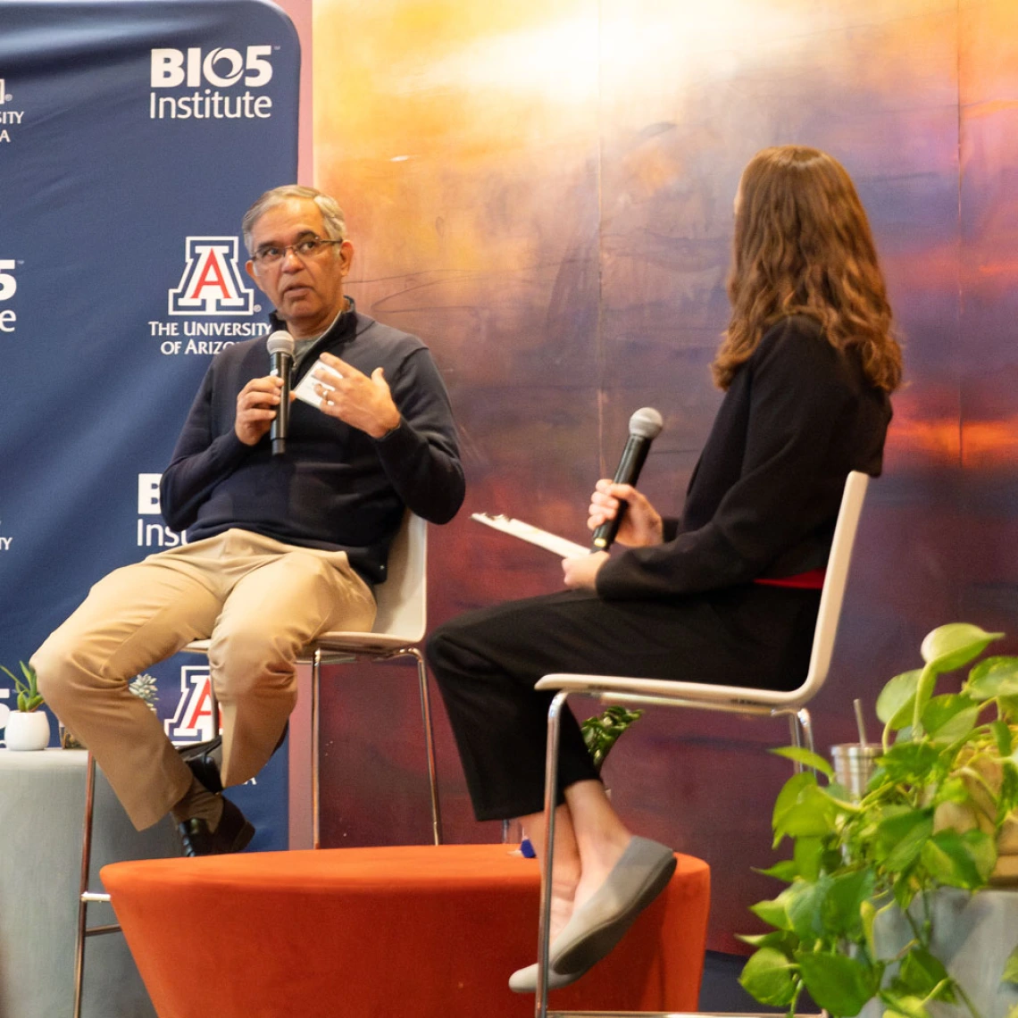 Two individuals seated on stage during a discussion at a BIO5 Institute event, University of Arizona. One person is gesturing while speaking and the other listens attentively. They are surrounded by banners with the BIO5 and University of Arizona logos.