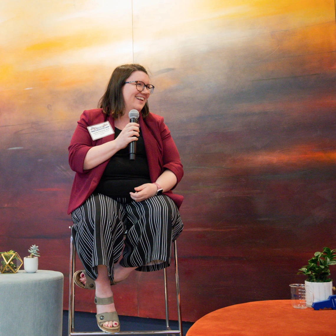 Person sitting on a stool, holding a microphone and speaking at an event, with a colorful abstract painting in the background.