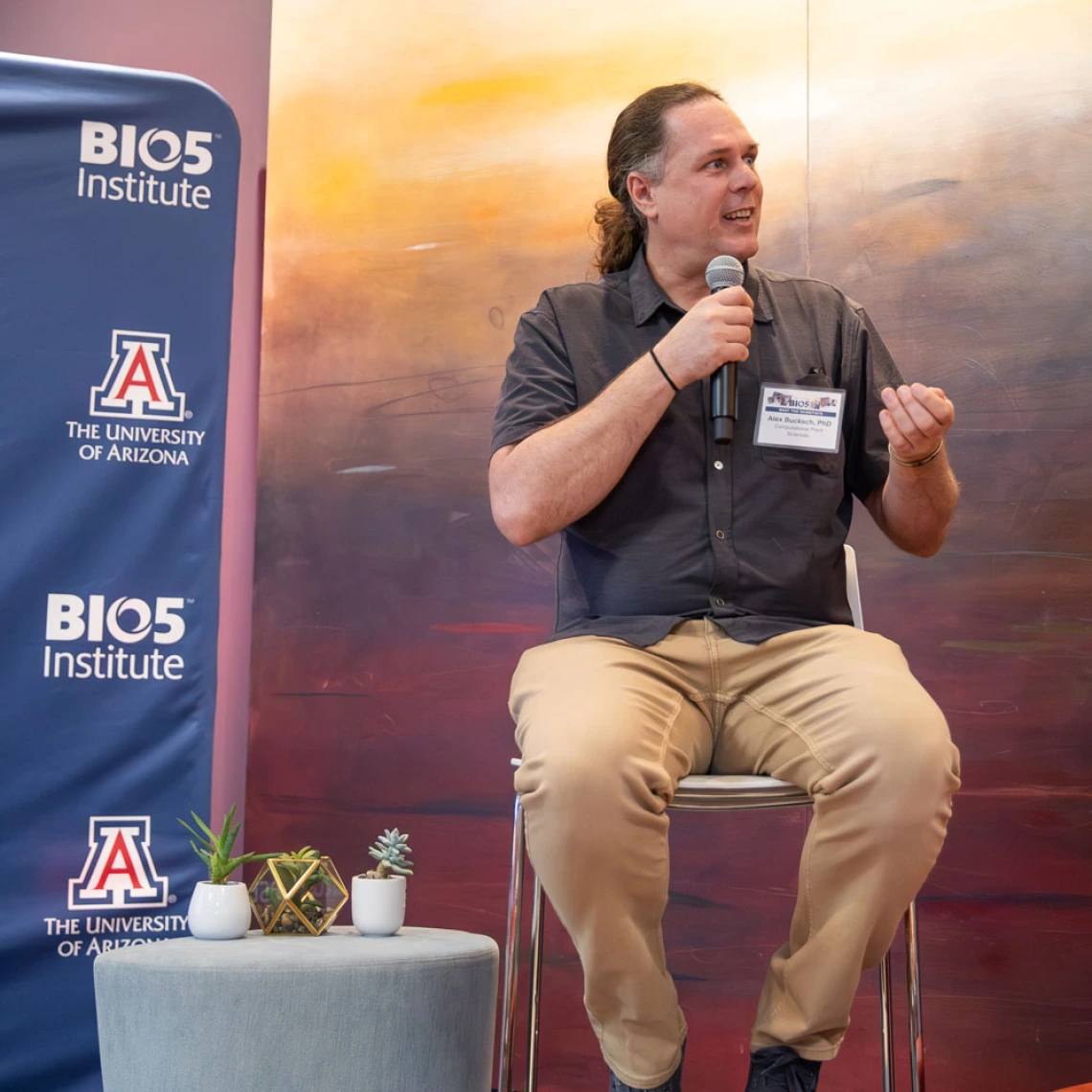 Person sitting on a stool, speaking into a microphone at a BIO5 Institute event, with a backdrop featuring a sunset landscape.