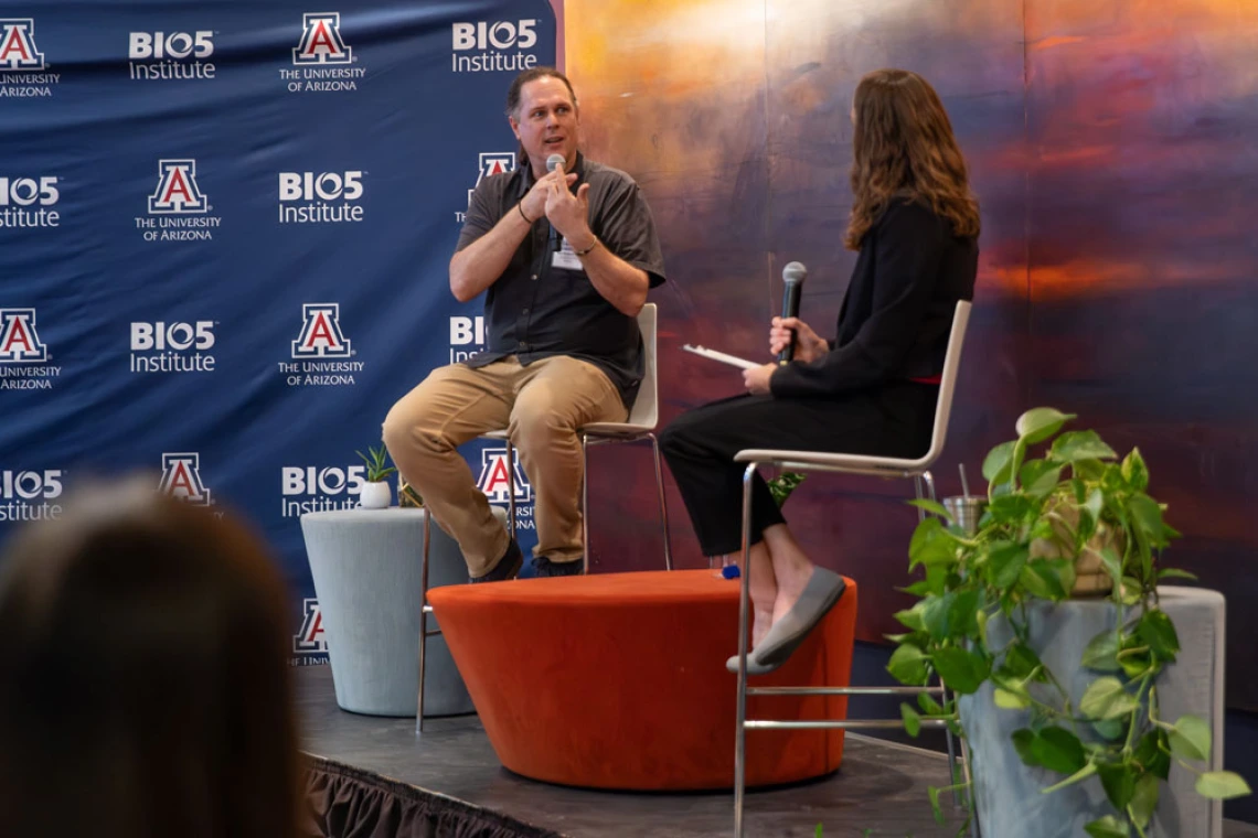 Two individuals participating in a discussion at the University of Arizona's BIO5 Institute. One is seated on a stool and speaking into a microphone, while the other listens attentively. The backdrop features the institute's logos and an abstract art piece.