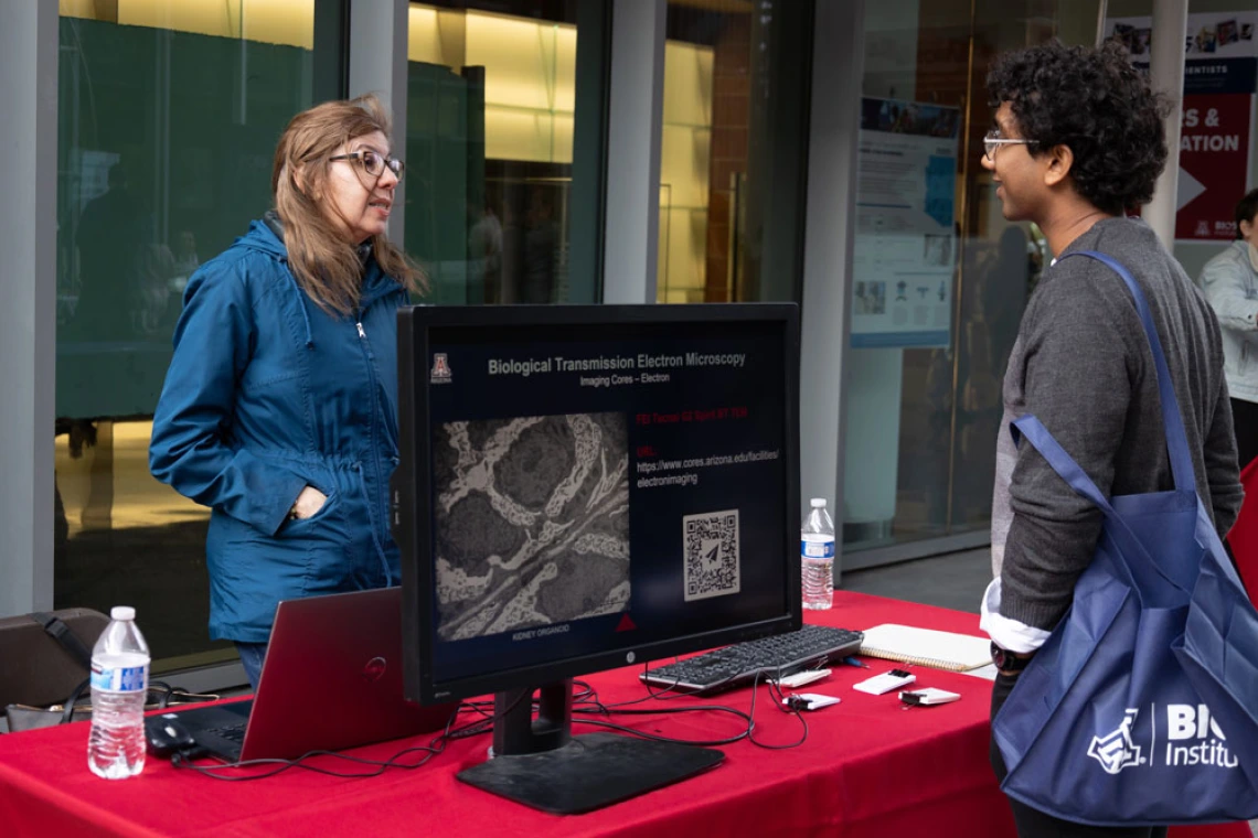 Two people are discussing at a booth with a sign labeled "Biological Transmission Electron Microscopy" at the BIO5 Institute. The monitor displays a microscopic image alongside QR codes.