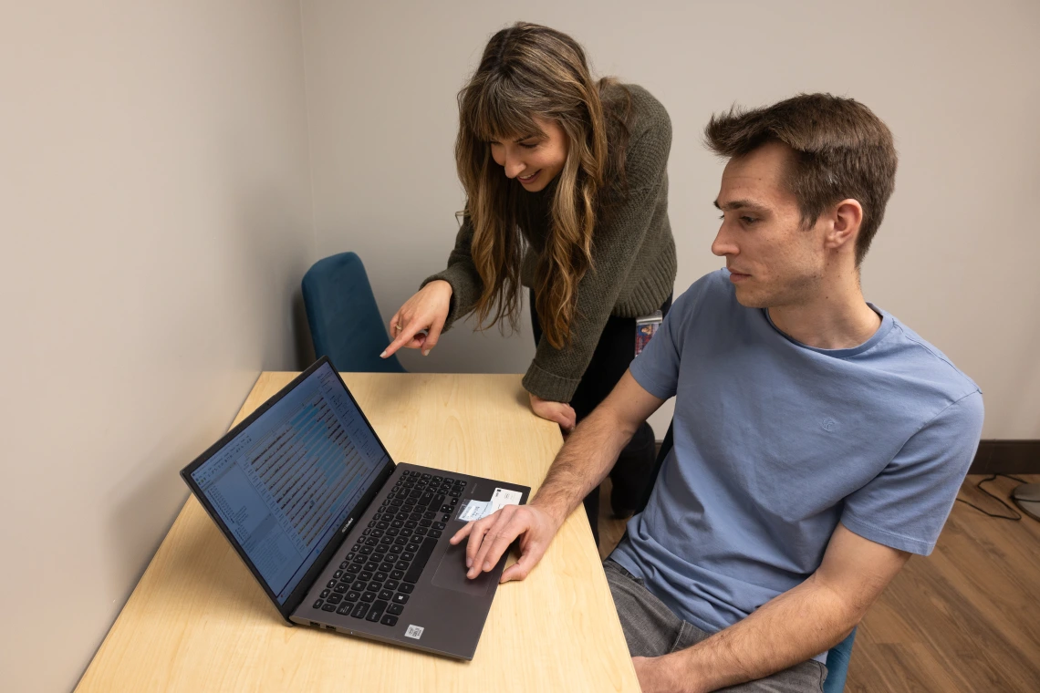 Two people, one pointing at a laptop screen and the other looking at it, are collaborating in an office setting. The laptop displays code or text.