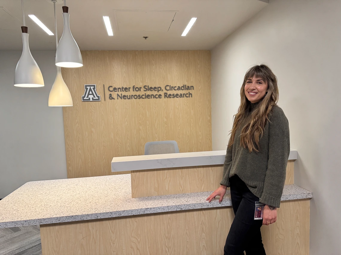 Person standing in a lobby at the Center for Sleep, Circadian & Neuroscience Research at the University of Arizona. The background features a reception desk with the center's logo.