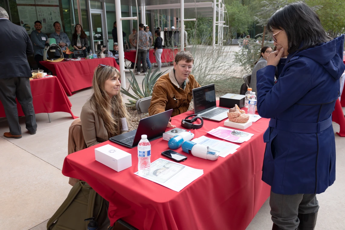 Two people sitting at a booth with a laptop and various items on a table, interacting with a standing visitor at an outdoor event. The booth has a red tablecloth and is set up near a building with glass doors and several onlookers in the background.