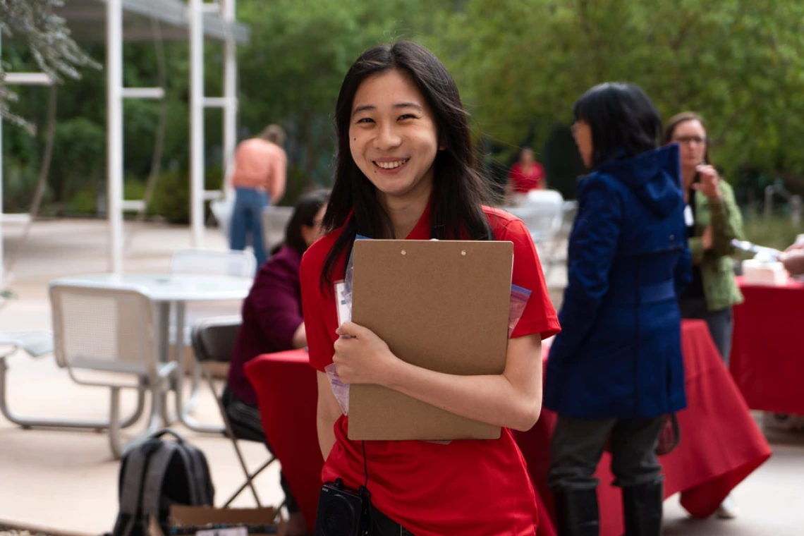 Young person in a red shirt smiling and holding a clip board with an event occuring in the background