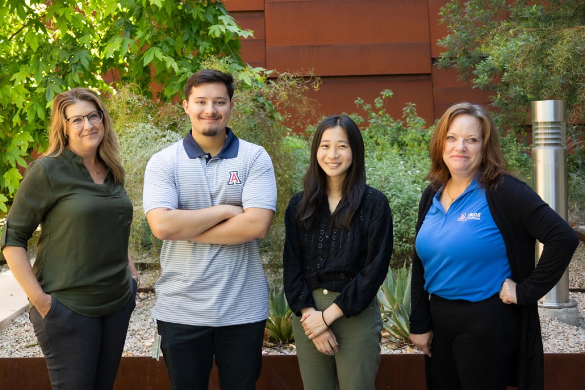 Four individuals standing outdoors in front of a building with lush greenery and a modern architectural background. Two are wearing university-branded apparel, indicating an affiliation with the University of Arizona.