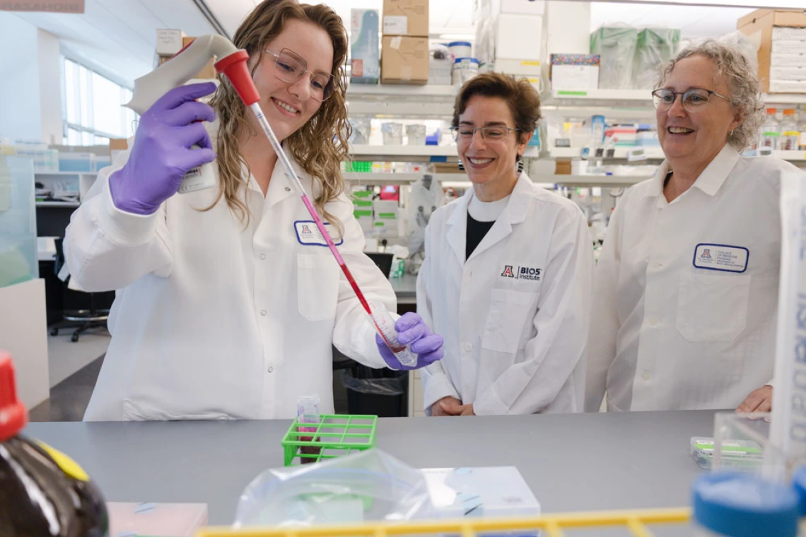 Three scientists in lab coats, one using a pipette, smiling in a laboratory setting.