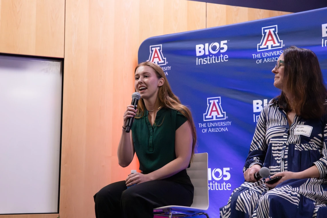 Person speaking into a microphone at a University of Arizona event, seated next to another participant in front of a banner with university logos.