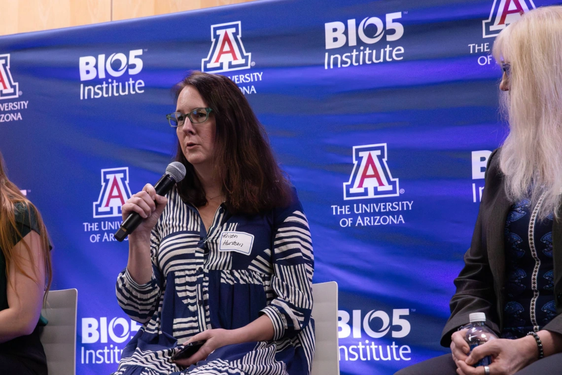 Person speaking into a microphone at a University of Arizona BIO5 Institute event, seated with others in front of a banner with the institute's logo.
