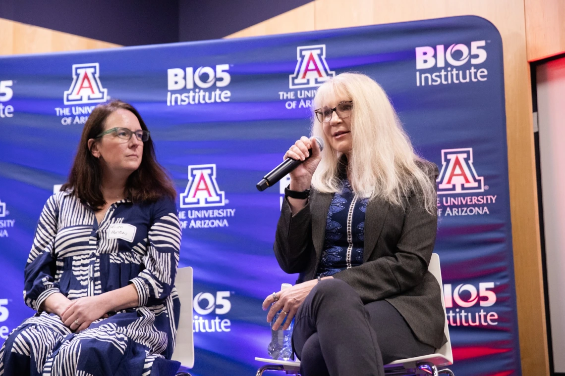 Two individuals seated on a stage at a BIO5 Institute event at the University of Arizona. One person is speaking into a microphone. They are both in front of a backdrop with the University of Arizona and BIO5 Institute logos.