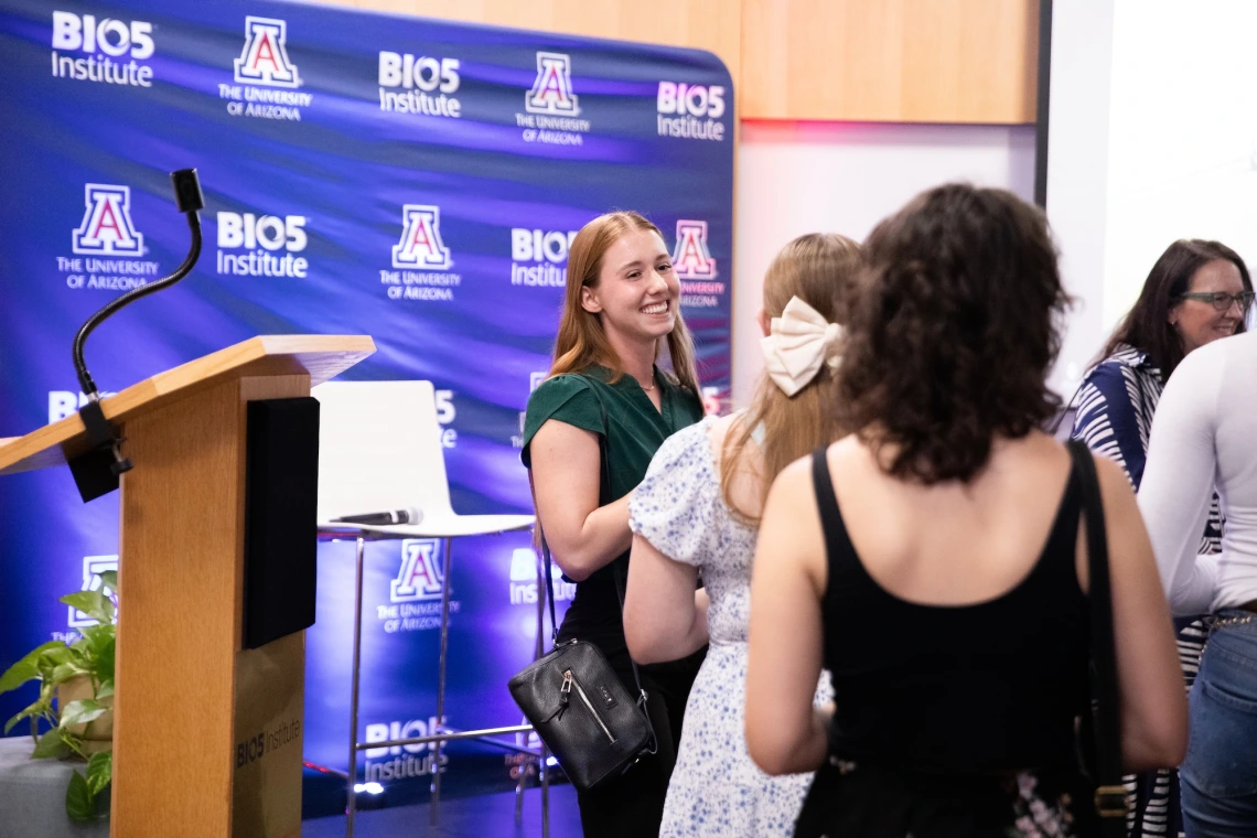 Person smiling and conversing with others at a University of Arizona BIO5 Institute event, with promotional banners in the background.