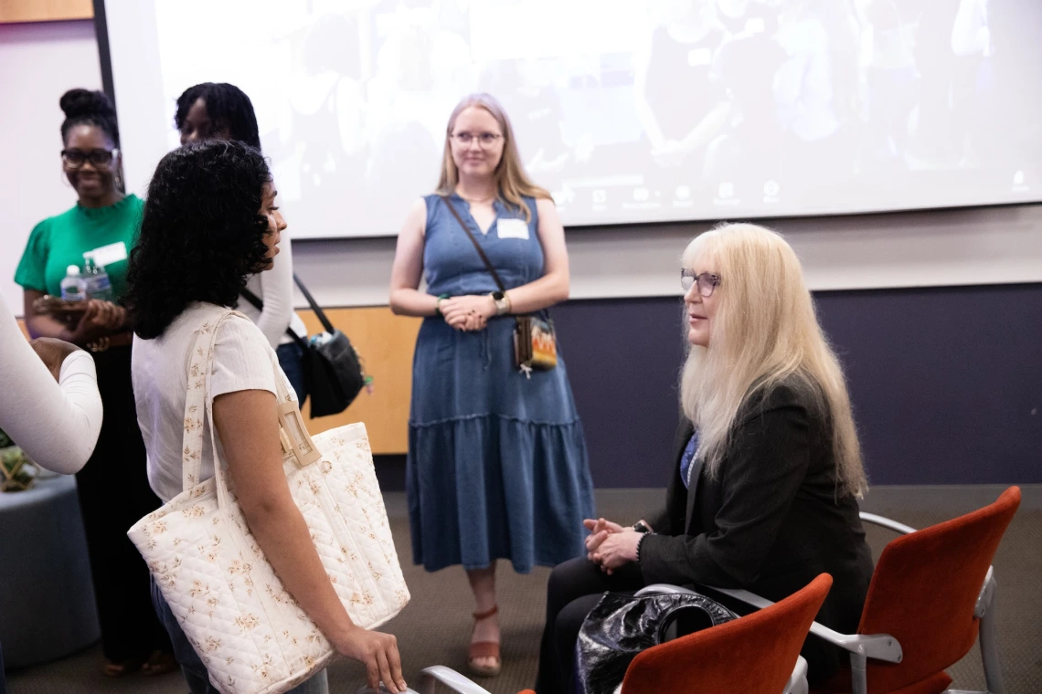 Group of individuals engaging in a conversation in a classroom setting with a large screen displaying additional participants in the background.