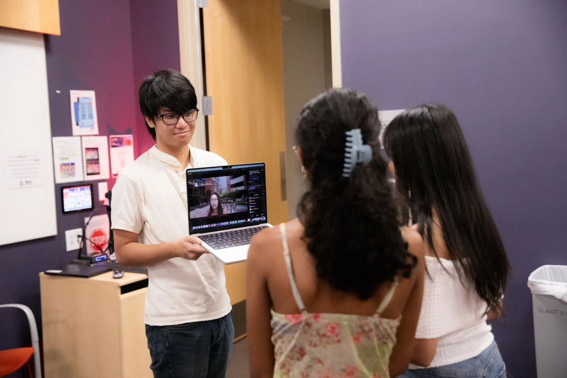 Three individuals in an indoor setting, one holding a laptop with a video call on screen, engaged in a discussion.