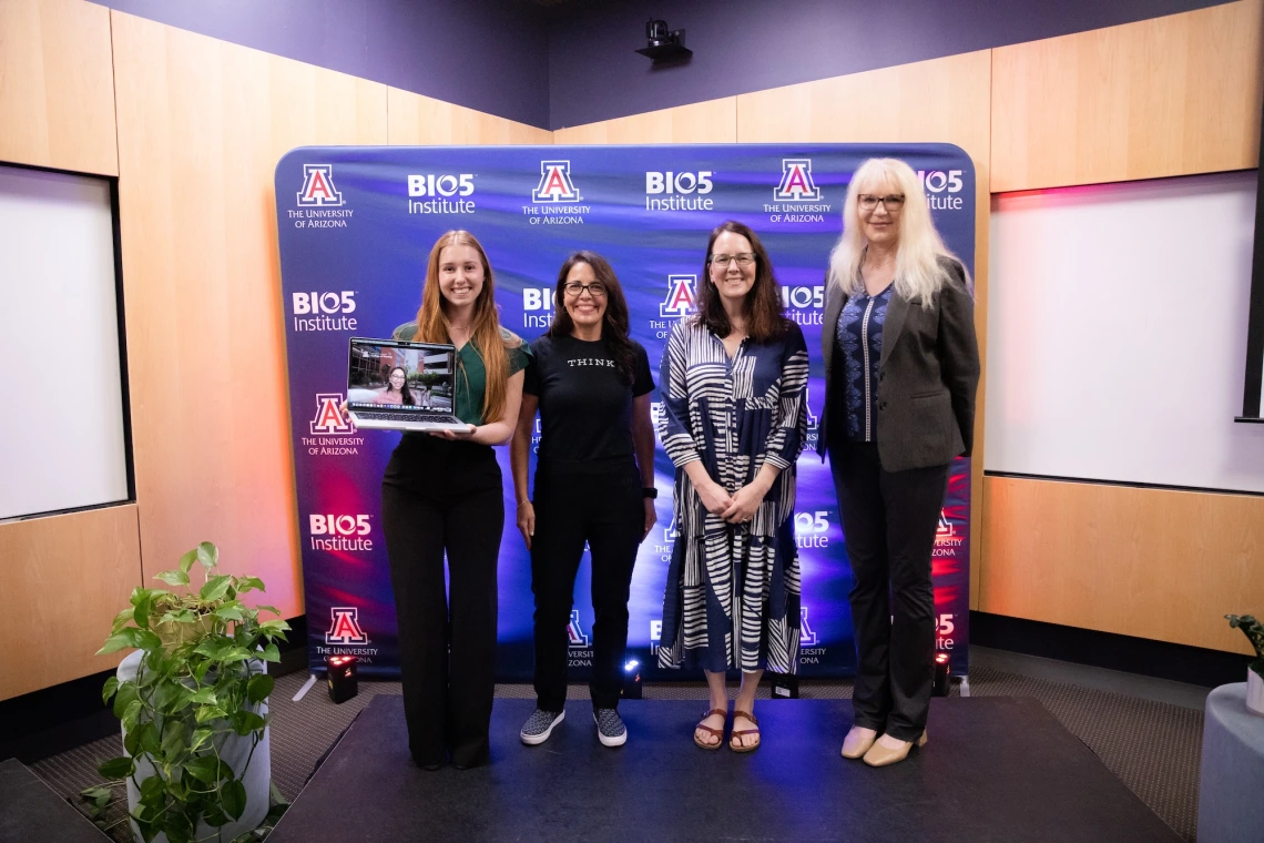 Four individuals smiling and posing together at the University of Arizona's BIO5 Institute, standing in front of a banner with the institute's logo. One person is holding a laptop.