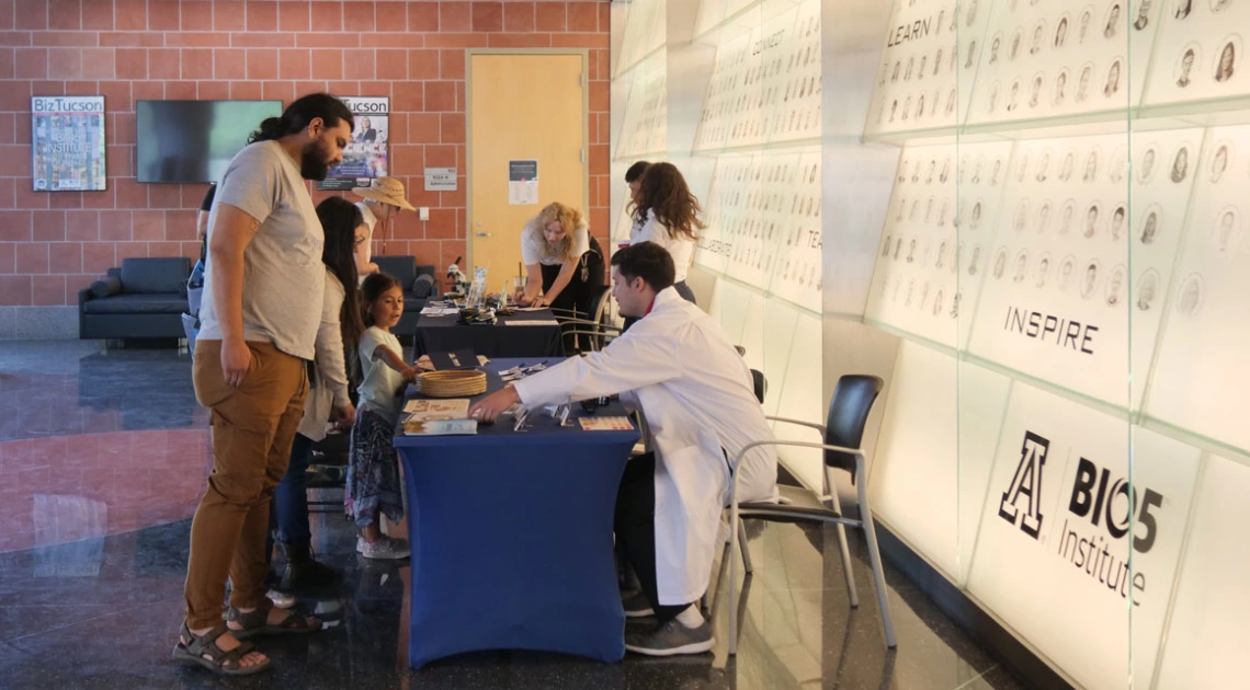 A person interacts with visitors at the Bios Institute booth inside a building with a wall featuring numerous etched portraits and inspirational words.